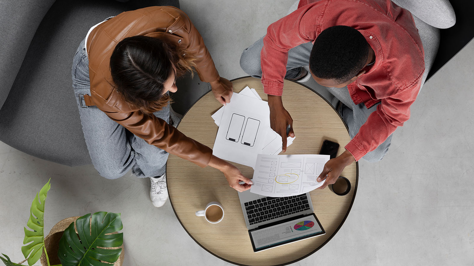 Two professionals collaborating at a wooden table on mobile app wireframes and UI mockups, with a laptop, smartphone, and coffee.