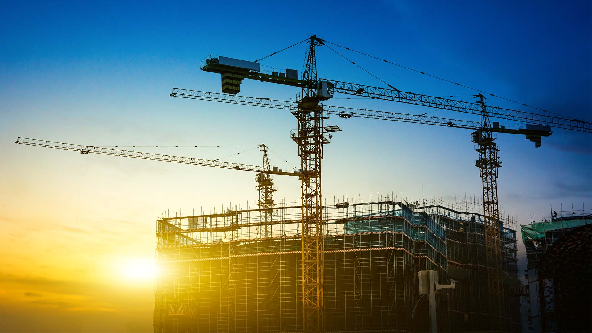 Sunrise construction site with multiple tower cranes working around a scaffolding-covered mid-construction building under a vibrant blue-to-orange sky, highlighting the scale of urban development and engineering.